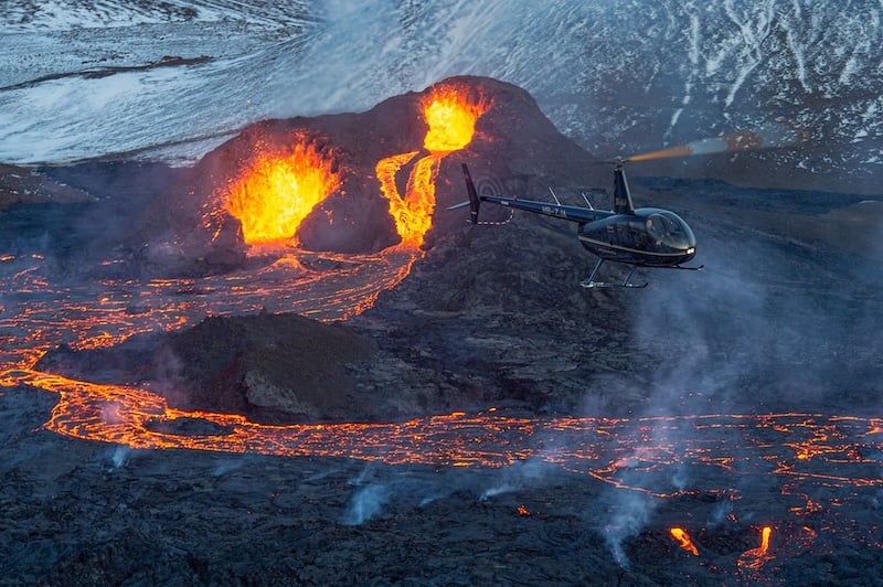 A Robinson R66 helicopter from Volcano Heli flying over an active volcanic eruption in Iceland, with glowing lava fountains and flowing lava fields visible below during a helicopter tour.
