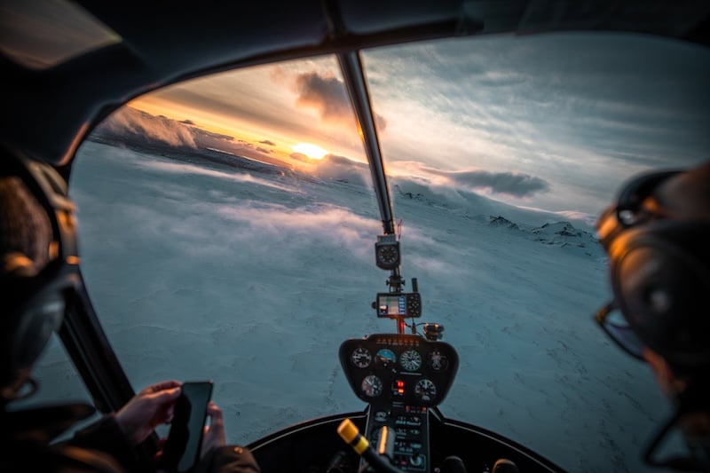 View from inside a Volcano Heli helicopter cockpit flying over a snow-covered glacier in Iceland at sunset, with the pilot visible and golden light breaking through clouds over mountain peaks.
