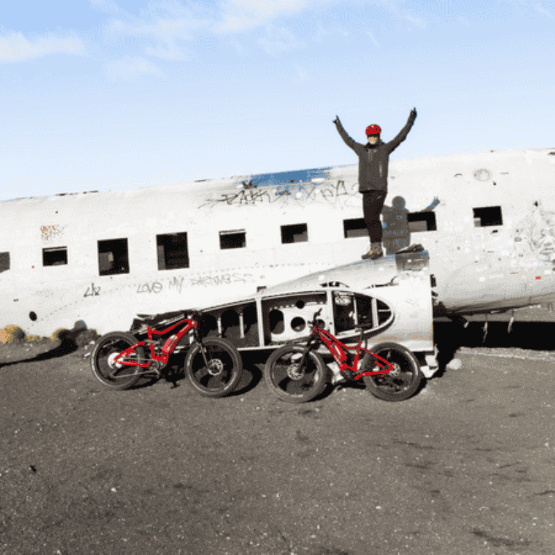 Two red electric fat bikes parked at the Sólheimasandur DC-3 plane wreck on Iceland's south coast, rider standing on top.