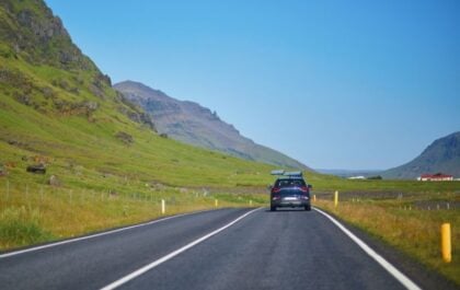 Rental SUV with a roof box driving along a paved road through green mountains on a sunny summer day in Iceland