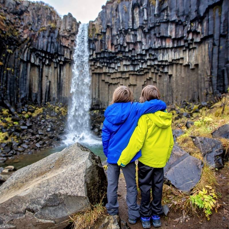 Two children in blue and green waterproof jackets standing arm-in-arm in front of Svartifoss waterfall in Skaftafell, southeast Iceland, surrounded by black basalt columns — a family-friendly hike in Iceland.