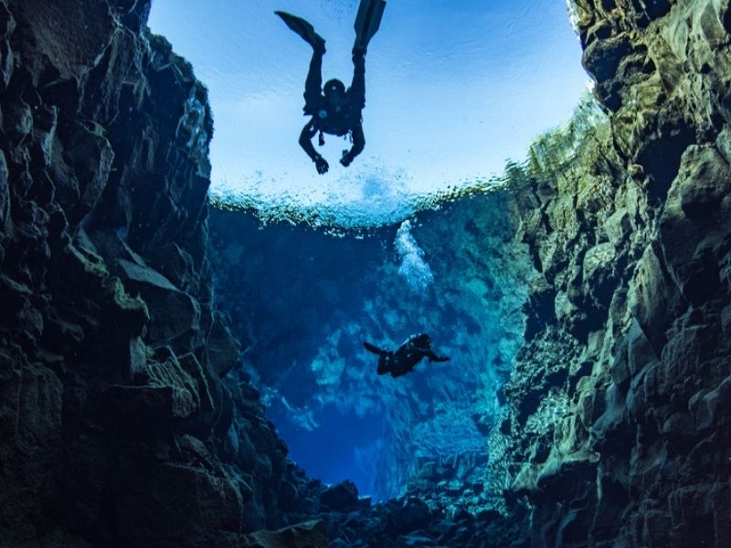 Two scuba divers descending into the deep, narrow Silfra fissure between towering lava rock walls in Þingvellir National Park