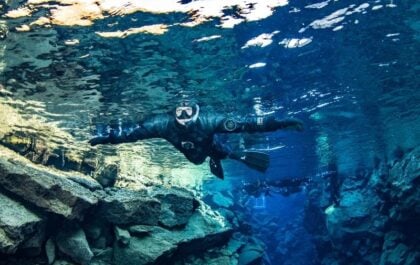 Snorkeler in a dry suit gliding through the clear blue water of the Silfra fissure, with lava rock walls below and other snorkelers visible further down the crack in Iceland