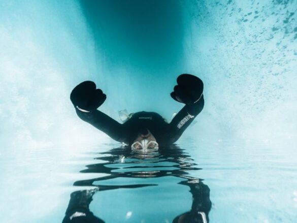 Freediver in a black wetsuit and gloves floating at the surface of a turquoise supraglacial lake, with the blue ice of Fjallsjökull glacier arching overhead and a mirror reflection on the still water.