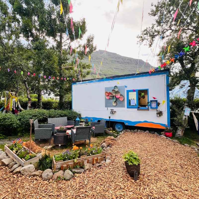 The La hamaca y la luna food truck in Seyðisfjörður, white with blue trim, in a garden with flower beds, garden seating, and colorful pinwheels strung in the trees, with a mountain rising behind.