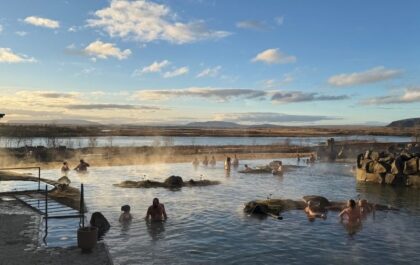 Bathers relaxing in the steaming geothermal water of Laugarás Lagoon in South Iceland, with moss-topped rock islets in the pool and the Hvítá river and distant mountains in the background under a partly cloudy sky.