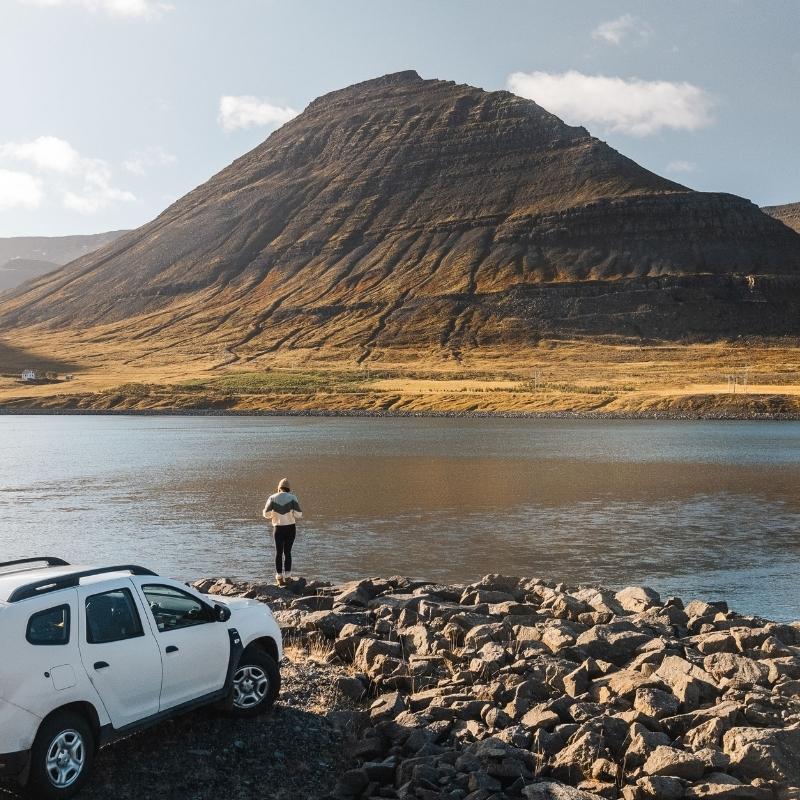 White Dacia Duster 4x4 rental car parked by a fjord in Iceland while a traveler stands on the rocky shore beneath a mountain. Photo credit: Go Car Rental