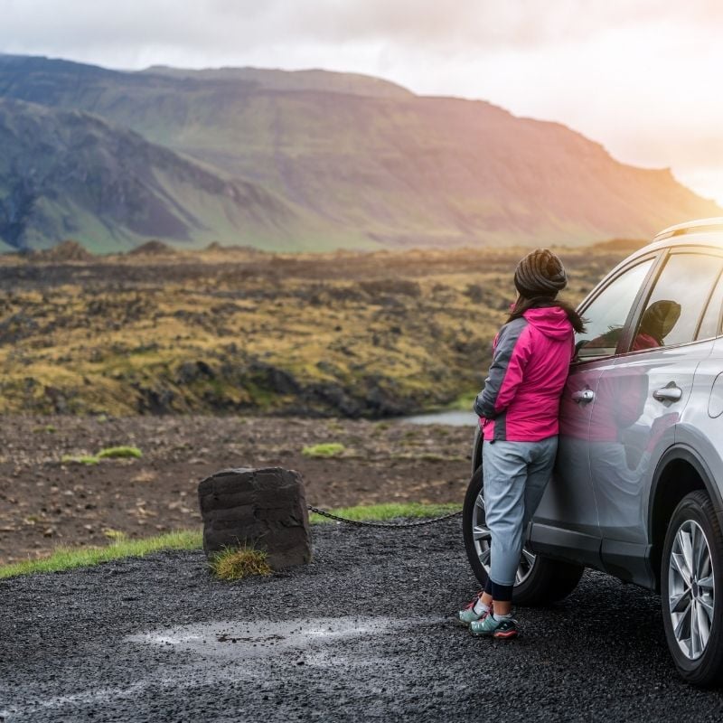 Traveler in a pink jacket leaning on a silver Toyota RAV4 rental car by a mossy lava field in Iceland, trunk open with luggage — a highland-permitted 4x4 from the Europcar Höldur fleet.