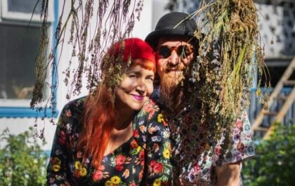 Tess Rivarola and Juanjo Ivaldi of La hamaca y la luna food truck, smiling at a wooden table in their Seyðisfjörður garden, surrounded by fresh peppers, apples, homemade drink in a swing-top bottle, and drying herbs hanging overhead.