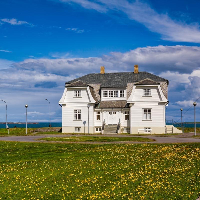 Höfði house, a white two-storey wooden villa with a slate roof, standing on a lawn of yellow dandelions beside the sea in Reykjavík, Iceland — one of the city's best-known landmarks and filming locations.