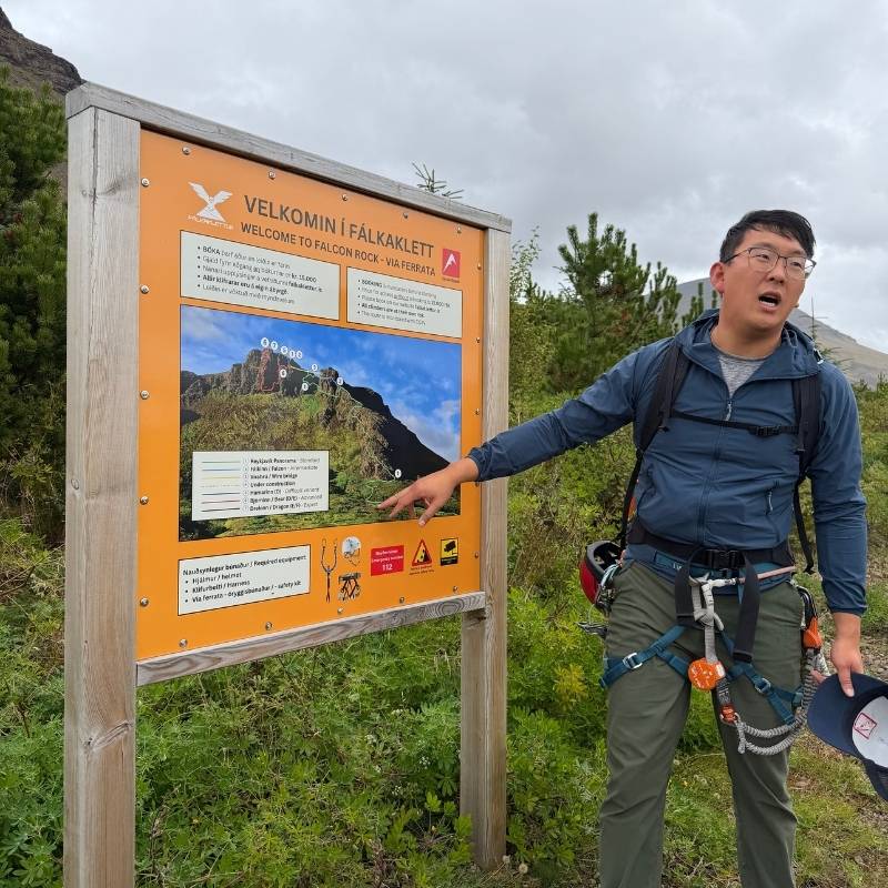 Guide Daniel Byung-chan Roh points at the Fálkaklettur via ferrata route map on an orange sign at the trailhead.