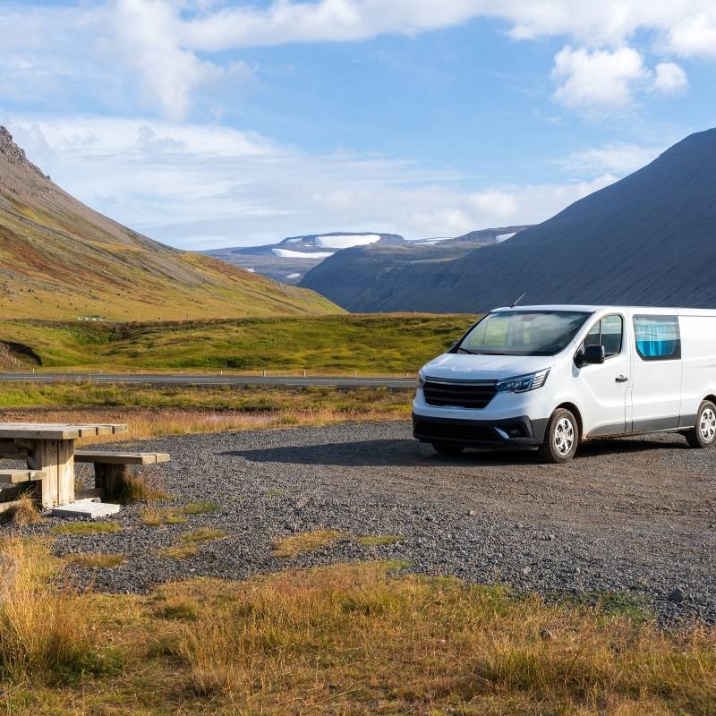 A white camper van parked on a gravel pull-off beside a picnic bench, below snow-streaked mountains in Iceland.
