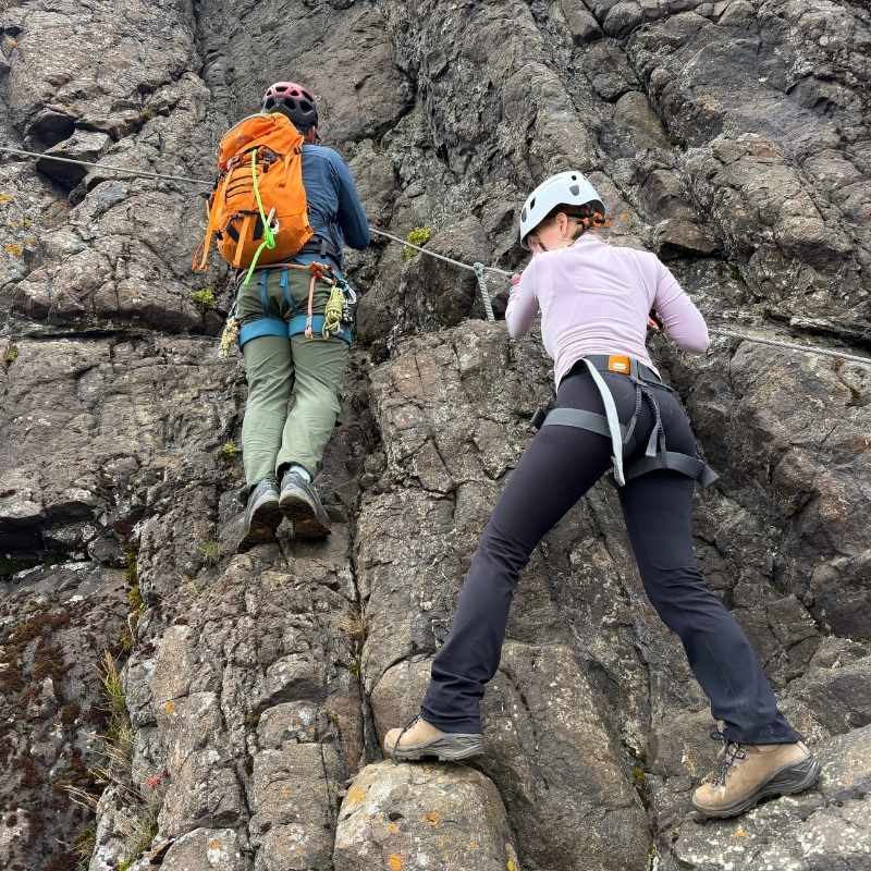 Two climbers on a steep section of the Fálkaklettur via ferrata, with the steel safety cable running across the rock beside them.