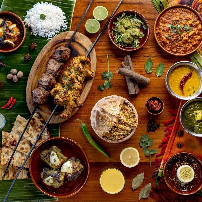 Overhead view of an Indian feast from Austur-Indía fjelagið in Reykjavík, laid out on banana leaves and a wooden table, with lamb and chicken tandoori skewers, garlic naan, biryani under a baked bread crust, saag paneer, yellow dal, a lamb shank curry, raita, a mango lassi and a round steel tin of colourful spices, surrounded by fresh herbs, limes, chillies and whole spices.